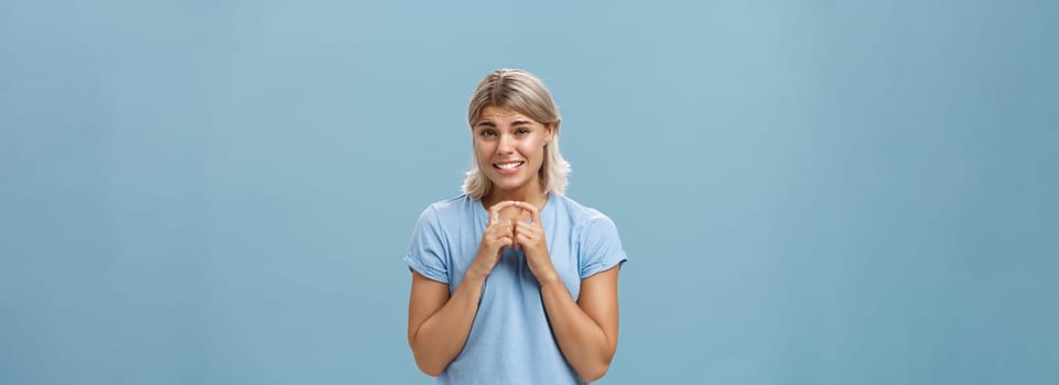Silly cute and worried blonde female friend with short haircut steepling index fingers and smiling with guilty sorry smile frowning while waiting for answer begging for help or favor over blue wall. Copy space