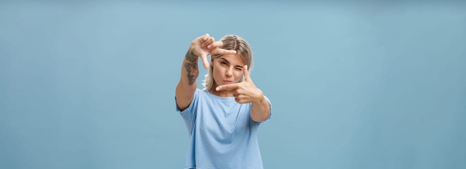 Artistic and creative female designer in stylish t-shirt closing one eye folding lip and making frame gesture while looking through it as if taking measurement or picturing something over blue wall. Copy space