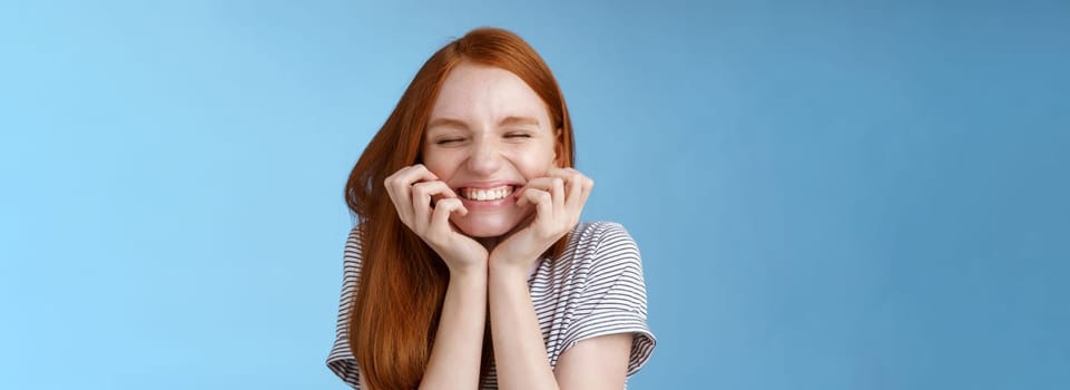 Cheerful carefree giggling ginger girl look happy bright close eyes smiling delighted hold hands cheeks having fun feeling excitement joy triumphing cheering good news, standing blue background.