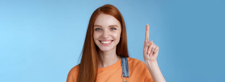 Confident happy charismatic ginger girl blue eyes freckles helpful showing product raising index finger up pointing cool copy space smiling broadly made choice recommend pick, studio background.