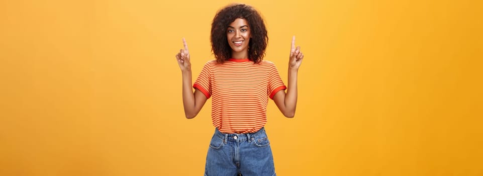 Looking only up and forward. Optimistic ambitious stylish dark-skinned female student in striped cool t-shirt and shorts raising hands pointing upwards and smiling friendly over orange wall.