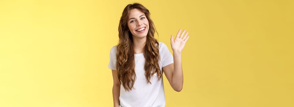 Modest cute friendly outgoing attractive european woman long curly haircut waving palm sociable conversation introduce herself smiling broadly say hi hello greeting coworker yellow background.