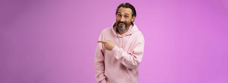 Friendly carefree caucasian bearded man in pink hoodie pointing right intoducing family members smiling proudly delighted showing blank copy space advertising, posing purple background.