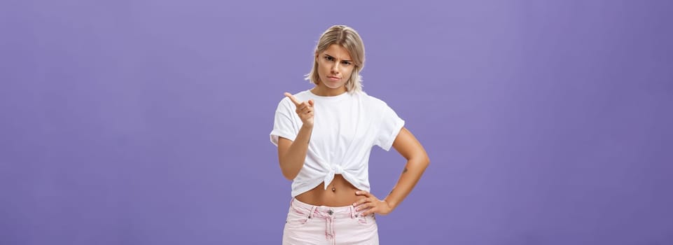 I told you behave properly. Strict and cute displeased blonde female in white t-shirt and shorts shaking index finger in strict pose looking from under forehead while scolding over blue background.