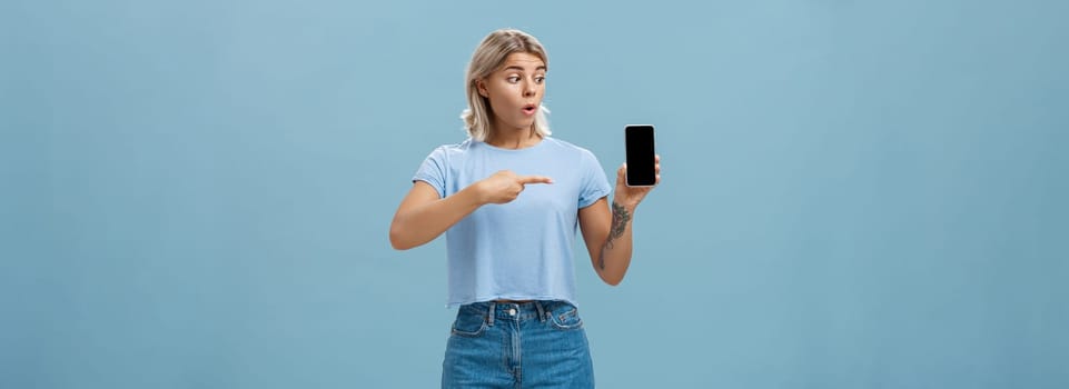 You should download app. Portrait of amazed shocked attractive blonde girl in trendy summer t-shirt holding smartphone gasping from surprise pointing at gadget screen posing over blue background.