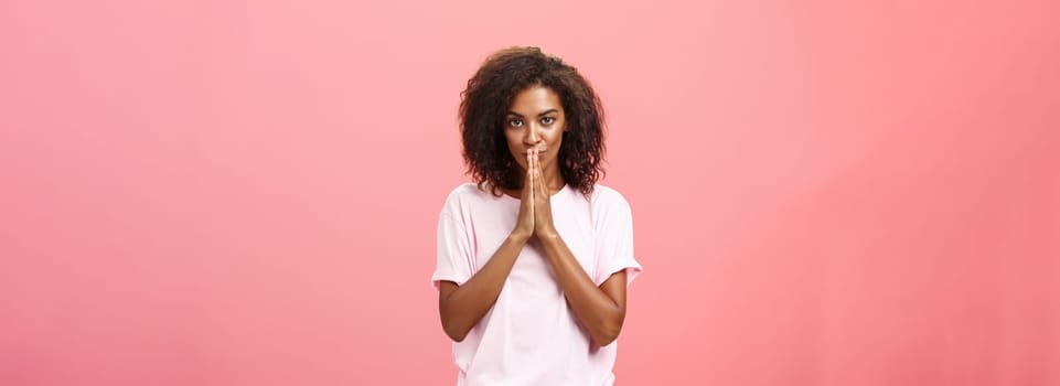 Creative and smart stylish attractive african american woman with curly hairstyle having great plan or idea feeling ambitious holding hands in pray over mouth gazing thoughtful at camera. Copy space