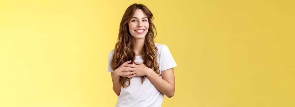 Lovely joyful tender feminine girlfriend long curly hairstyle touch heart feel heartbeat delighted receive lovely heartwarming gift smiling broadly satisfied flirty gazing camera yellow background.