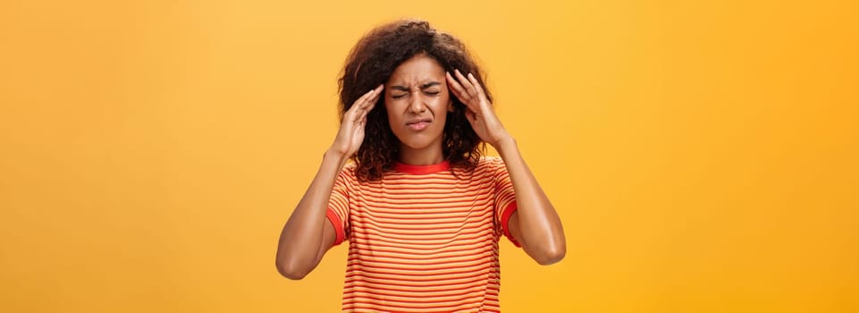 Woman cannot recall important information looking intense trying concetrate under pressure feeling tensed touching temples closing eyes while thinking hard posing against orange background.