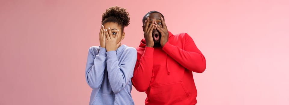 Funny cute carefree loving african american boyfriend girlfriend fool around hide faces palms peeking through fingers mimicking joking around make funny mimics, standing pink background surprised.