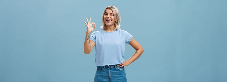 Confident and smart girl got everything under control. Portrait of optimistic good-looking girl with fair hair and tanned skin winking showing okay or perfect gesture standing over blue wall.