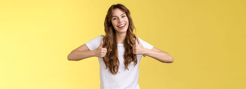 Outgoing positive confident lively young girl long curly haircut support great effor show thumbs up positive reply approving good choice like your outfit smiling broadly yellow background. Lifestyle.