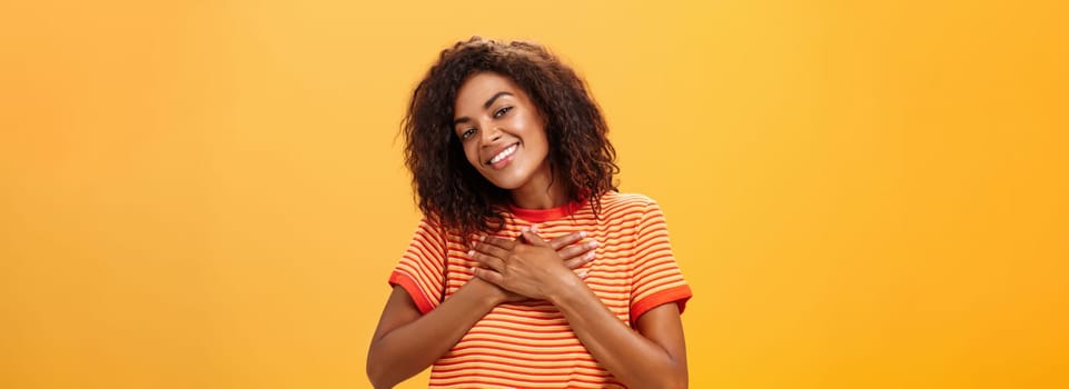 Waist-up shot of charming sensual dark-skinned woman with curly hairstyle in trendy t-shirt holding palms on heart looking with pleased loving smile feeling grateful and delighted over orange wall. Lifestyle.