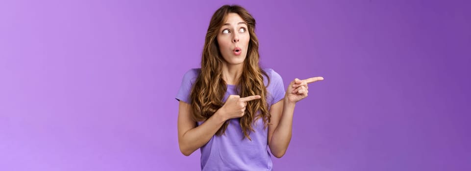 Curious surprised cute curly-haired caucasian female coworker discuss project amazed awesome cool result turn pointing left impressed folding lips wow tempating try new product purple background. Lifestyle.