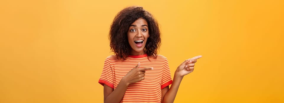 Portrait of amazed excited charismatic dark-skinned young pretty girl with afro hairstyle in trendy striped t-shirt pointing left delighted and fascinated posing against orange background. Lifestyle.