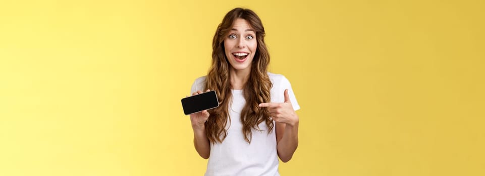 Cheerful surprised cute lucky girl beat best score awesome game showing smartphone display pointing index finger horizontal screen mobile phone introduce cool app smiling broadly yellow background. Lifestyle.