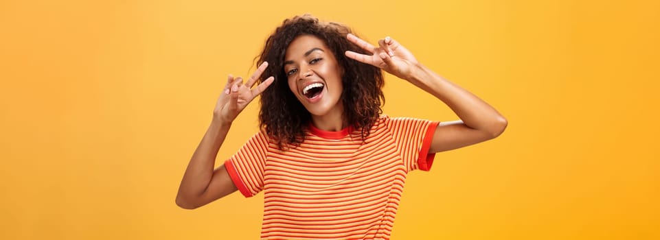 Nobody gonna spoil my perfect mood. Portrait of happy optimistic attractive dark-skinned female model with curly hairstyle tilting head smiling carefree showing peace or victory signs near face. Copy space