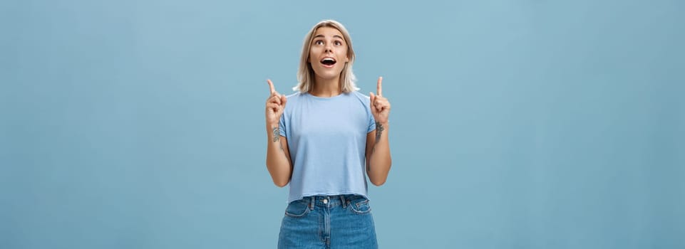 Lifestyle. Indoor shot of impressed speechless attractive fair-haired female student in casual t-shirt and denim shorts dropping jaw from amazement pointing and looking up intrigued over blue wall.