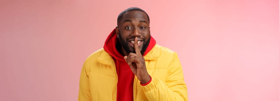 Lifestyle. Charming happy young african-american bearded guy in yellow jacket red hoodie prepare lovely birthday surprise smiling show shush shh gesture index finger on mouth promise keep secret.