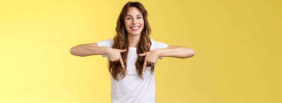 Lively helpful good-looking caucasian girl curly long hair showing you excellent place find hairdresser pointing down index fingers smiling toothy happy delighted grin stand yellow background. Lifestyle.