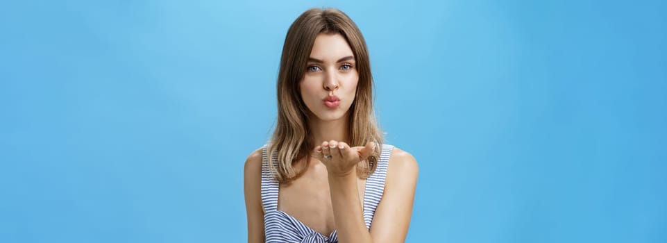 Charming confident and flirty girlfriend with chestnut hair and natural makeup folding lips holding palm near mouth sending air kiss at camera sensually expressing love and romance over blue wall. Lifestyle.
