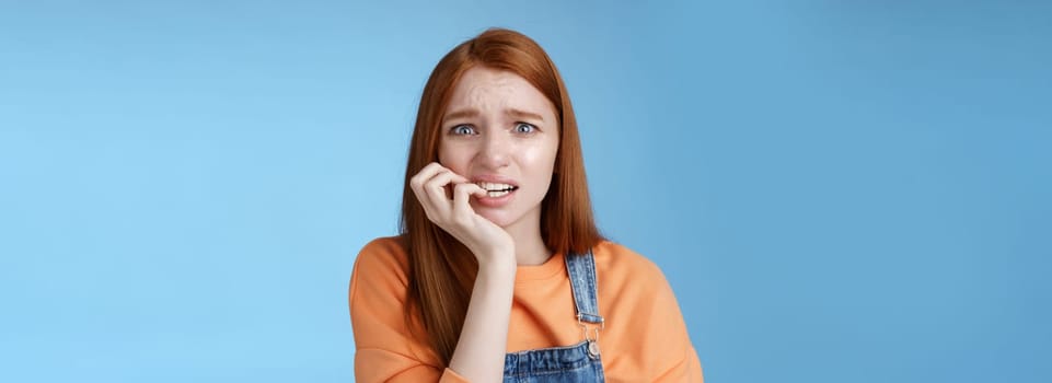 Intense worried scared unsure young redhead panicking silly girl frowning look upset anxious biting fingernails emotional frightened fired standing blue background emotional terrified. Copy space