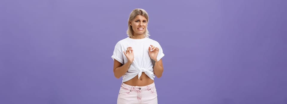 Ew take disgusting thing away from me. Stylish feminine good-looking blond woman in cropped top with tanned skin and pierced belly looking at creepy think with dislike and aversion over purple wall.