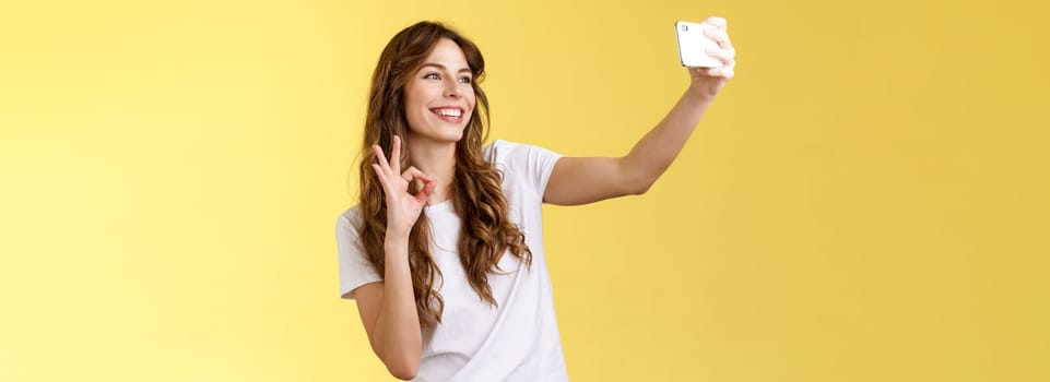 Stylish carefree urban confident curly-haired girl recording video blog followers extend arm hold smartphone taking selfie front camera show okay ok approval gesture yellow background. Lifestyle.