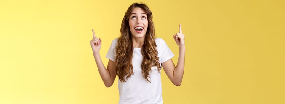 Happy impressed attractive girl long curly haircut drop jaw amused smiling surprised delighted look pointing up index fingers stare admiration fascinated awesome promo offer yellow background.Lifestyle.