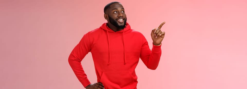 Happy charismatic delighted african-american bearded guy smiling white teeth look pointing intrigued upper left corner curious what interesting product standing impressed pink background.