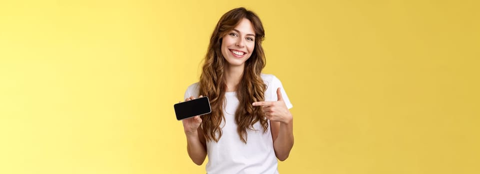 Cheerful good-looking friendly carefree young girl hold smartphone horizontal pointing index finger mobile phone screen smiling broadly introduce awesome game app stand yellow background.