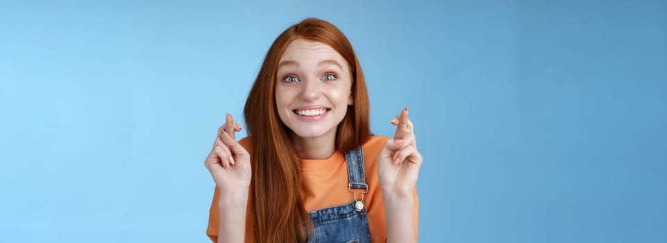 Excited emotional happy cheerful redhead girl smiling optimistic stare surprised thrilled cross fingers good luck believe dream come true make wish anticipating only good news, blue background.