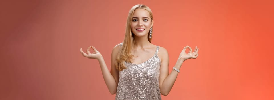 Lifestyle. Calm relieved happy fabulous young blond woman in silver stylish luxury dress standing relaxed red background zen lotus pose reach nirvana smiling peaceful carefree meditating breathing practice.