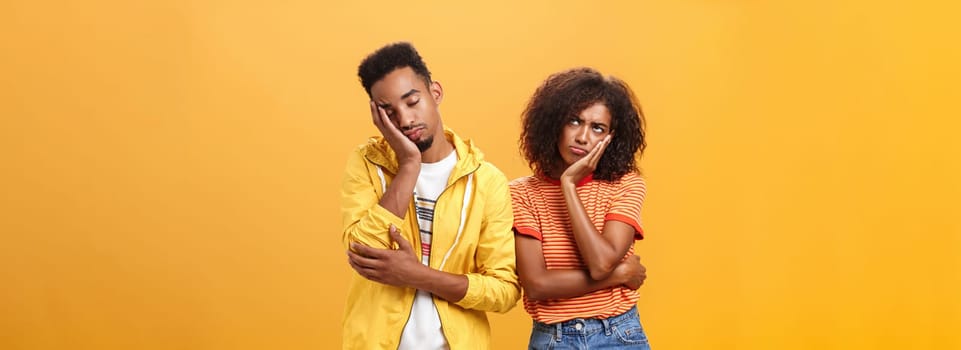 Girl feeling displeased and offended on boyfriend who fell asleep during date pursing lips frowning looking up while boyfriend leaning head on face with closed eyes and tired look over orange wall. Lifestyle.