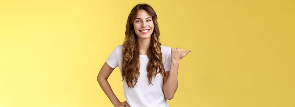 Friendly outgoing cheerful european cute female giving tips showing direction lively smiling toothy happily have nice pleasant conversation pointing left thumb introduce promo yellow background.