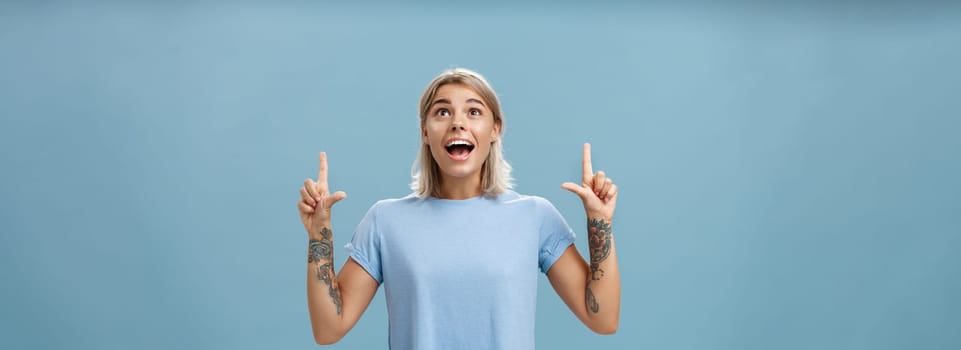 Studio shot of impressed speechless happy good-looking woman with tattoos on arms dropping jaw from amazement and joy gazing fascinated and pointing up standing over blue background. Emotions concept