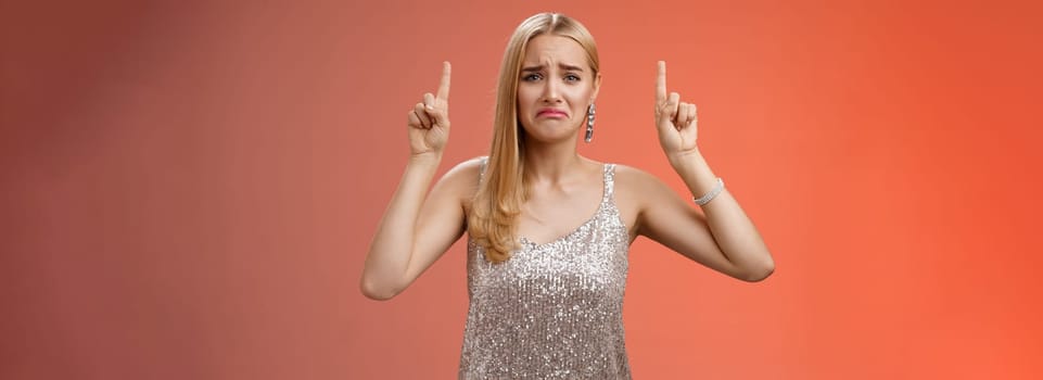 Upset whining immature spoiled blond rich girl in silver glittering dress pouting frowning gonna cry pointing up regret jealousy begging buy expensive shoes, standing red background displeased.