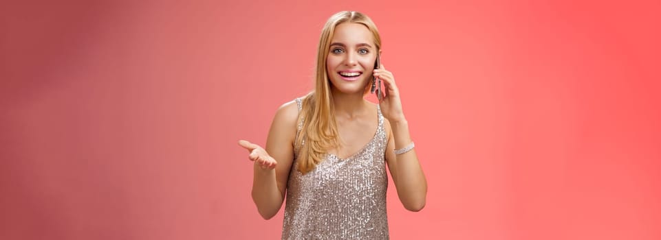 Joyful talkative outgoing attractive blond woman talking friend smartphone gesturing amused smiling broadly retelling fresh rumors after party wearing silver stylish dress, red background.