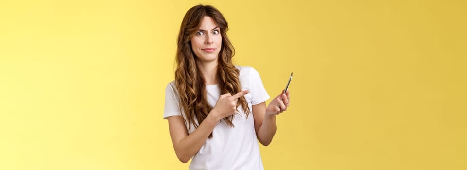Girl answers strange weird call receive crazy upsetting message cringe doubtful displeased smirking dismay pointing suspicious smartphone stand yellow background intense frustrated.