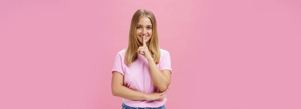 Shh keep it secret. Portrait of charismatic cheerful chubby girl with tanned skin and fair hair showing shush gesture with index finger over mouth smiling hiding surprise posing over pink background.