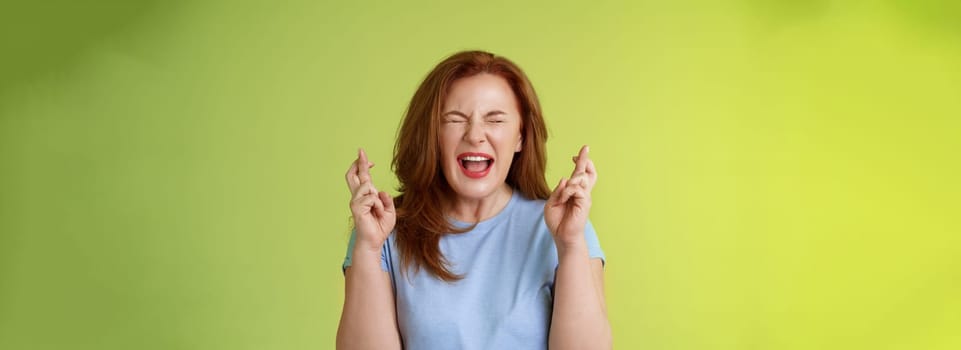 Woman wants win badly. Enthusiastic lucky redhead middle-aged 50s female pleading implore god make dream come true cross fingers good luck wishing closed eyes open mouth excitement green background.