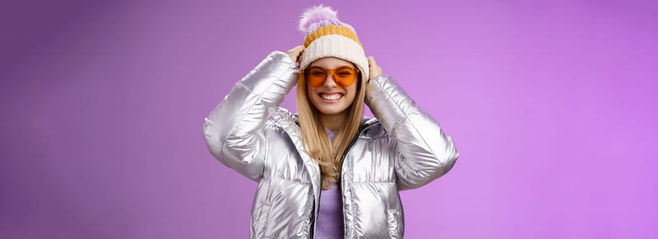 Energized daring sassy young attractive woman having fun friends winter trip learn snowboarding smiling cheeky enjoying vacation put-on hat wearing silver warm jacket sunglasses, purple background.