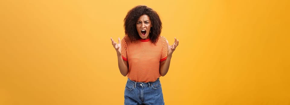 Pissed and outraged angry african american young female in stylish outfit raising palms gesturing and yelling losing temper arguing with cheating partner feeling fed up over orange background. Lifestyle.