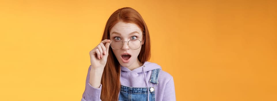 Shocked impressed redhead coworker hearing last intimate rumors drop jaw staring full disbelief camera confused touch glasses seem unsure truth lie standing orange background astonished.