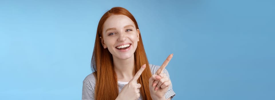 Happy carefree smiling redhead caucasian girl ginger straight hairstyle pointing upper left corner laughing joyfully amused cool promo giving recommendation try new product, standing blue background.