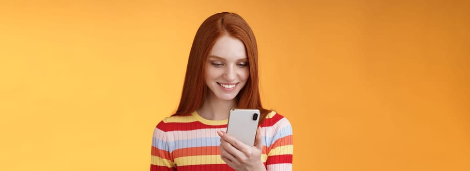 Charming modern redhead girl college student checking message box holding smartphone look happy smiling delighted cellphone display receive hundred likes photo post online, orange background.