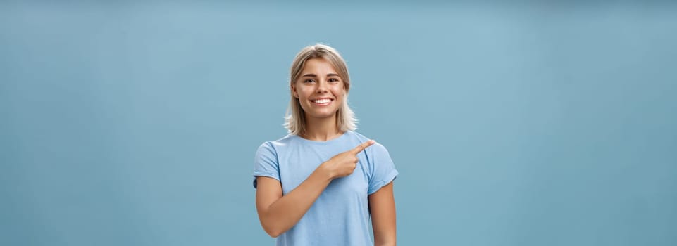 Come with me. Portrait of stylish pleased good-looking female student with blond hair in trendy t-shirt pointing at upper left corner and smiling with delight and happiness posing over blue background.