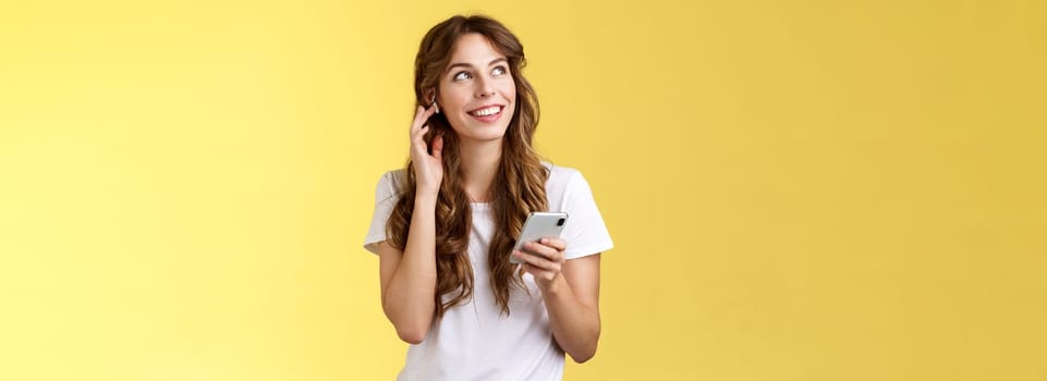 Dreamy happy cheerful curly-haired girl look around contemplate beautiful summer weather listening music touch wireless earbud calling friend talking via earphones hold smartphone yellow background. Lifestyle.