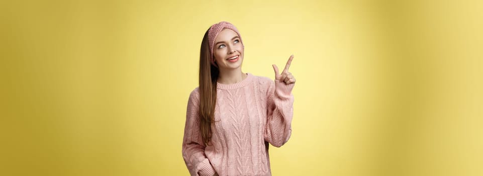 Portrait of amused interested cute 20s european girl wearing sweater, headband looking upper left corner pointing sideways smiling intrigued, ecstatic, captured by curious promo over yellow wall.