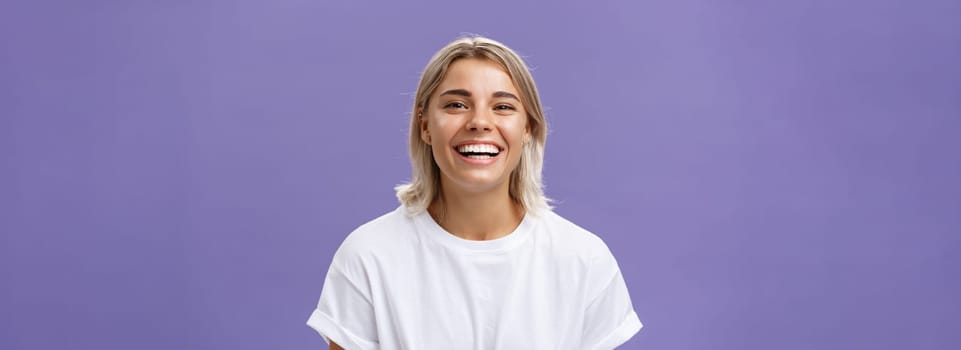 Close-up shot of joyful charming blonde female with delighted and pleased smile standing in white t-shirt over purple background spending time in awesome amusing company. Emotions concept