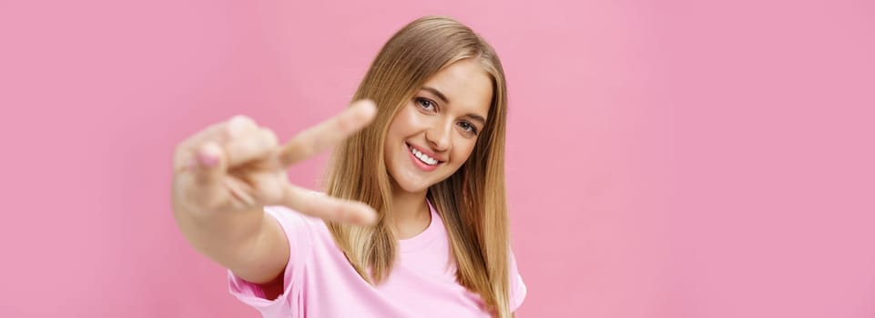 Peaceful and joyful good-looking young girl with straight blond hair in t-shirt pulling hand with peace sign towards camera, tilting head smiling friendly posing against pink background.